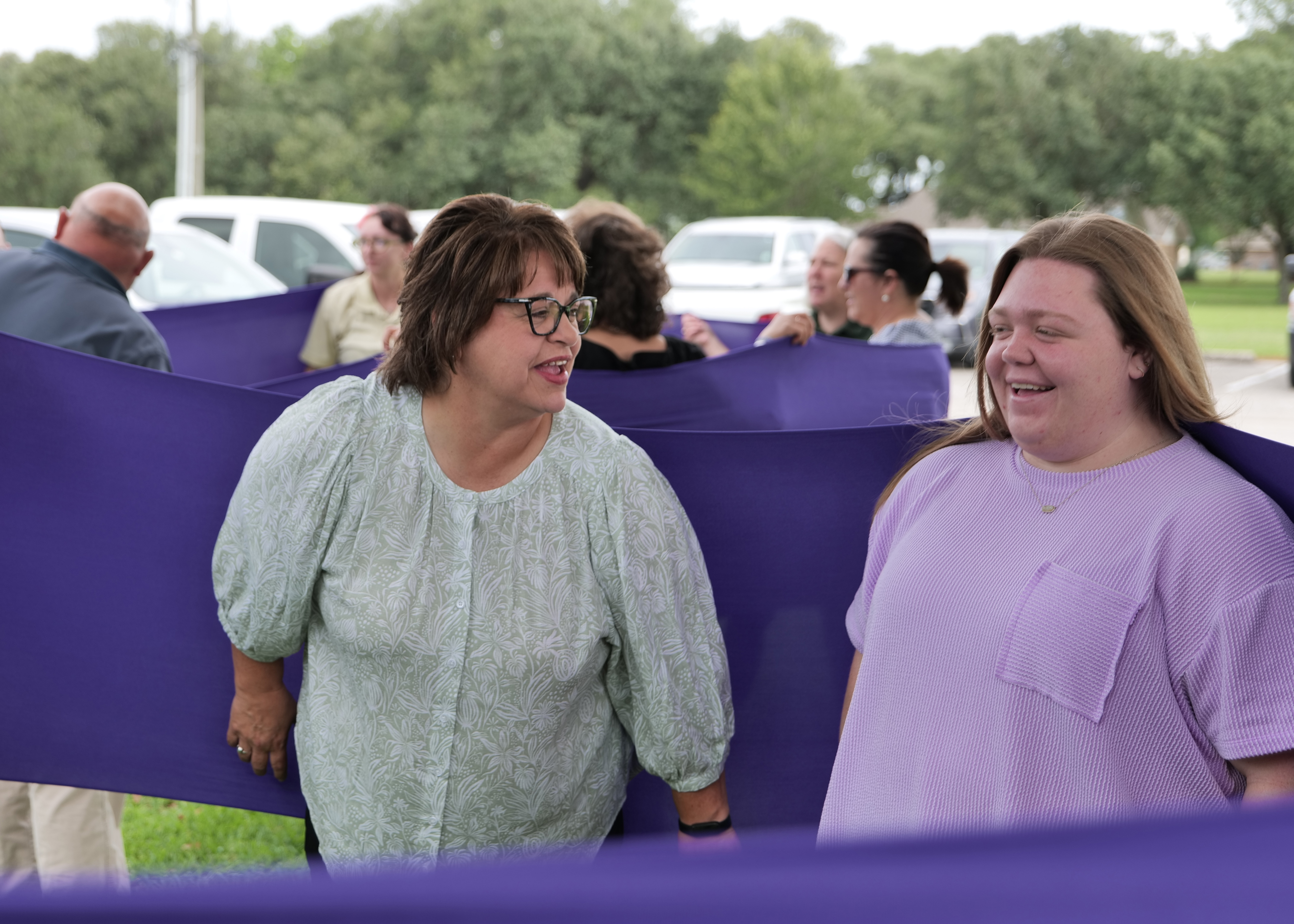 Two women smile and talk while standing against purple fabric. 